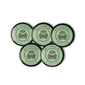 Puede incluir: Cinco botones redondos con un dise&ntilde;o verde y negro. Cada bot&oacute;n presenta una rana de dibujos animados con una corona, la palabra "RESIST" y el texto "ANTIFASCIST SOCIAL CLUB".