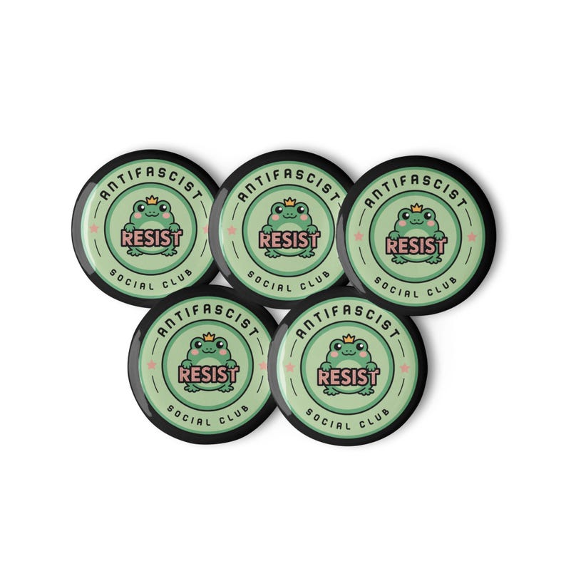 Puede incluir: Cinco botones redondos con un dise&ntilde;o verde y negro. Cada bot&oacute;n presenta una rana de dibujos animados con una corona, la palabra "RESIST" y el texto "ANTIFASCIST SOCIAL CLUB".