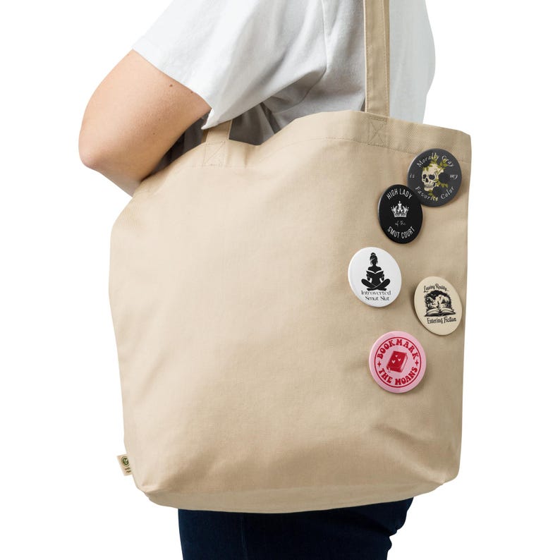 May include: A beige canvas tote bag with several round pinback buttons attached. The buttons feature various designs, including a skull, a seated figure, and text. The bag is held by a person wearing a white shirt.