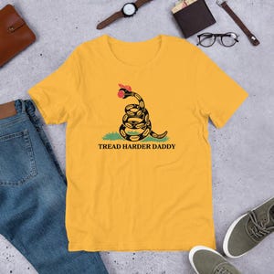 May include: Mustard yellow t-shirt with a coiled snake graphic, red tongue, and the text "TREAD HARDER DADDY". The shirt is made of a soft material and is suitable for casual wear.