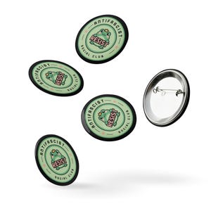 Puede incluir: Botones negros con un fondo verde claro y una ilustraci&oacute;n de una rana de dibujos animados con una corona. El texto "RESIST" est&aacute; en el centro, con "ANTIFASCIST SOCIAL CLUB" alrededor del borde. Se muestra un bot&oacute;n desde atr&aacute;s.