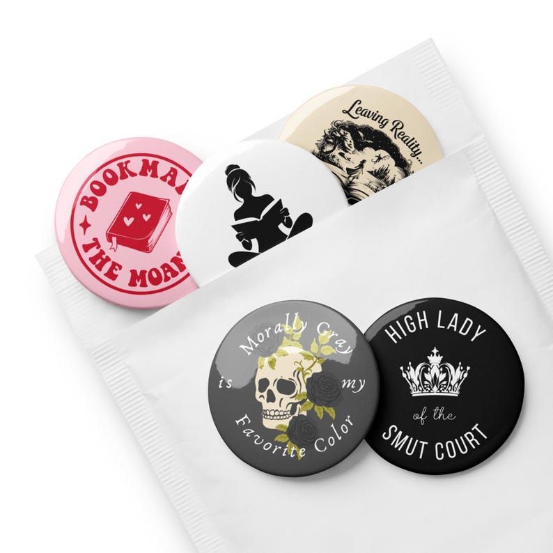 May include: A collection of round pinback buttons with various designs and text. One pink button reads "BOOK THE MOAN" with a book illustration. Other buttons feature a skull, a silhouette of a person reading, and the text "HIGH LADY of the SMUT COURT".