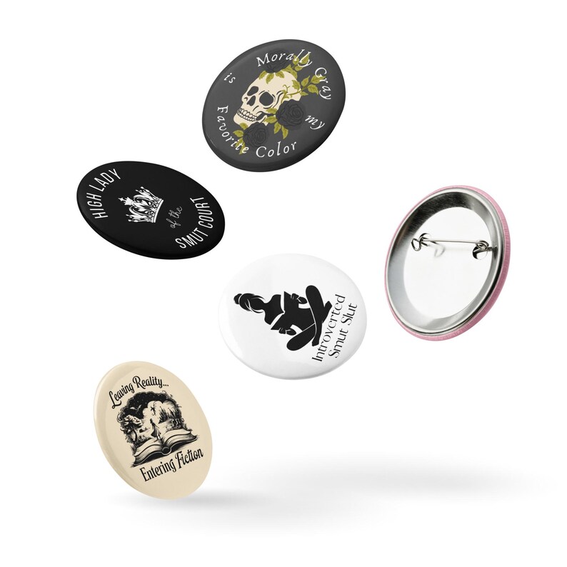 May include: A collection of five round and oval buttons with various designs and text. One button features a skull with floral accents and the text "Morally Gray is my Favorite Color." Other buttons include text like "High Lady of the Smut Court" and "Introverted Smut Slut."