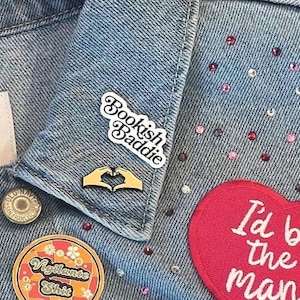 Könnte beinhalten: Nahaufnahme einer hellblauen Jeansjacke, verziert mit verschiedenen Ansteckern und Aufnähern. Die Jacke hat einen "Bookish Baddie" Anstecker, einen herzförmigen Handgesten-Anstecker und einen runden "Vigilante Shit" Anstecker. Ein roter Herzaufnäher liest "I'd be the man".