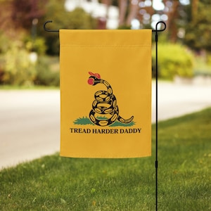 May include: Yellow garden flag featuring a coiled snake wearing a red hat and a green patch of grass. The flag reads "Tread Harder Daddy" in black text. The flag is hanging on a black metal stand in a grassy yard.