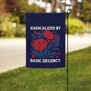 Op de afbeelding: Een marineblauwe tuinvaandel met de tekst "RADICALIZED BY BASIC DECENCY" in het wit. De vlag heeft een bloemenontwerp met rode bloemen en blauwe bladeren. De vlag hangt aan een zwarte metalen paal in een tuin.