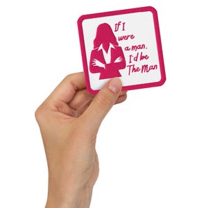 May include: A square, white patch with a pink border and the text "If I were a man, I'd be The Man." A pink silhouette of a person in a business suit is also on the patch.