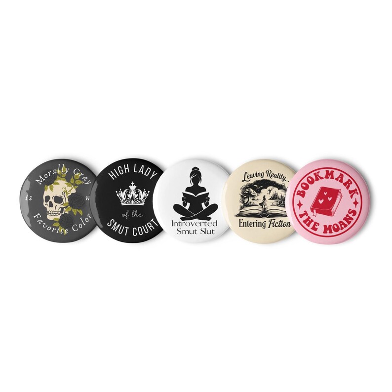 May include: A collection of five round pinback buttons. The buttons feature various designs and text, including a skull, a crown, a silhouette of a person reading, an open book, and a book with a heart. The color palette includes black, white, gray, and pink.