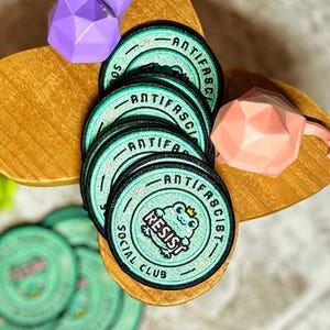May include: A stack of teal and black embroidered patches with the words "ANTIFASCIST SOCIAL CLUB" and a cartoon frog wearing a crown. The patches are on a wooden surface with geometric paper sculptures.