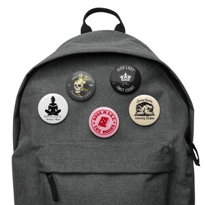 May include: A gray backpack with five round pinback buttons attached. The buttons feature various designs, including text and illustrations. One button reads "Morally Gray is my Favorite Color."