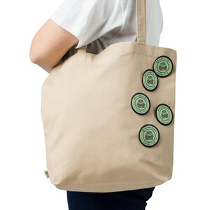 Puede incluir: Bolso tote de lona beige con varios parches redondos. Los parches son verdes y llevan las palabras "ANTIFASCIST" y "RESIST" en un dise&ntilde;o circular. El bolso tote tiene un dise&ntilde;o sencillo y lo sostiene una persona.