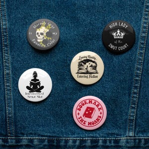 May include: A collection of round buttons with various designs and text. One button features a skull and roses, another says "High Lady of the Smut Court," and others have book-related themes. The buttons are on a denim background.