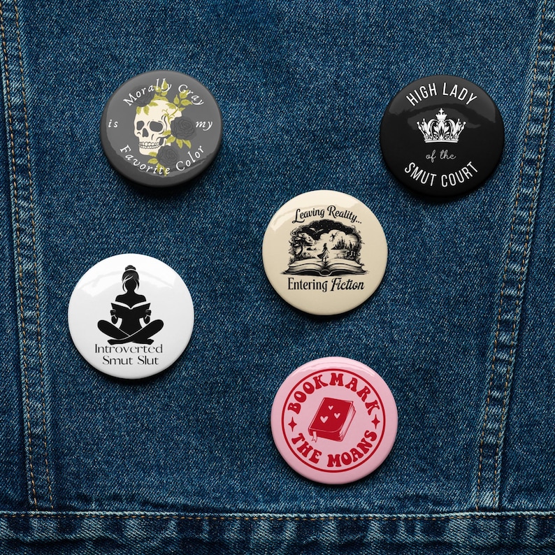 May include: A collection of round buttons with various designs and text. One button features a skull and roses, another says "High Lady of the Smut Court," and others have book-related themes. The buttons are on a denim background.
