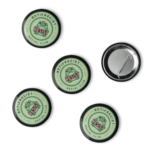 Puede incluir: Se muestran cinco insignias redondas. Cuatro insignias presentan una rana verde claro con una corona, la palabra "RESIST" y el texto "ANTIFASCIST SOCIAL CLUB" en un dise&ntilde;o circular. Una insignia muestra la parte posterior con el alfiler.