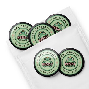 Puede incluir: Un conjunto de botones redondos negros y verdes con las palabras "ANTIFASCIST" y "RESIST". Cada bot&oacute;n presenta una rana de dibujos animados con una corona. Los botones est&aacute;n empaquetados en una funda de papel blanco. Las palabras "SOCIAL CLUB" tambi&eacute;n est&aacute;n presentes.