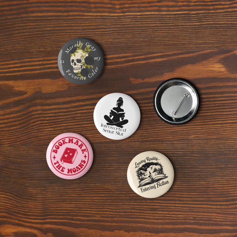 May include: A collection of round pinback buttons with various designs and text. One button features a skull and the words "Morally Gray is my Favorite Color." Other buttons have phrases like "Introverted Smut Slut" and "Leaving Reality... Entering Fiction."
