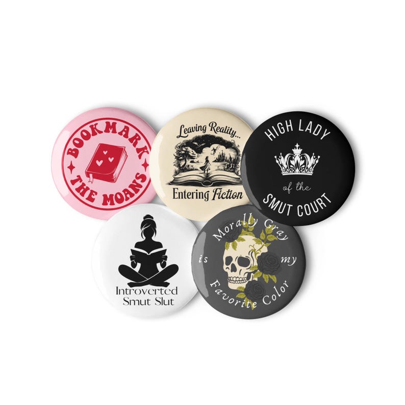 May include: A collection of five round buttons with various designs and text. The buttons feature phrases like "Bookmark the Moans," "Leaving Reality... Entering Fiction," and "High Lady of the Smut Court." Other buttons include a skull and a silhouette of a person reading.