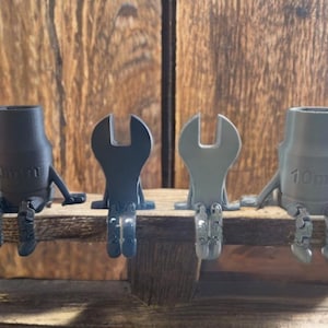 Può includere: Quattro statuette stampate in 3D raffiguranti chiavi a bussola e chiavi inglesi in varie tonalità di grigio e nero, ciascuna con braccia e gambe, sedute su una superficie di legno. Le chiavi a bussola hanno la scritta "10mm".