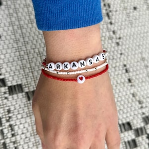 May include: Three beaded bracelets stacked on a wrist. The top bracelet spells "ARKANSAS" in white letter beads. The middle bracelet is white with silver beads. The bottom bracelet is red with a heart bead.