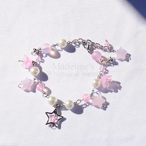 May include: A delicate bracelet featuring pink star and flower charms, pearl beads, and pink beads strung on a silver chain. A silver star charm hangs from the bracelet. The text "Madeline's Whimsical Wares" is visible.