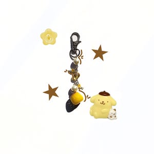 May include: A yellow keychain with a silver clasp, adorned with pearls, yellow beads, and a fluffy yellow pom-pom. A yellow cartoon character with a brown beret and a small white cat are also featured.