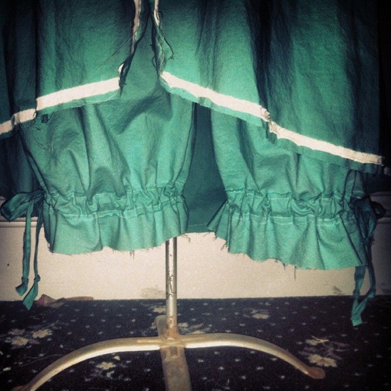 May include: A green fabric skirt with a white trim and a gathered bottom. The skirt is on a metal stand.