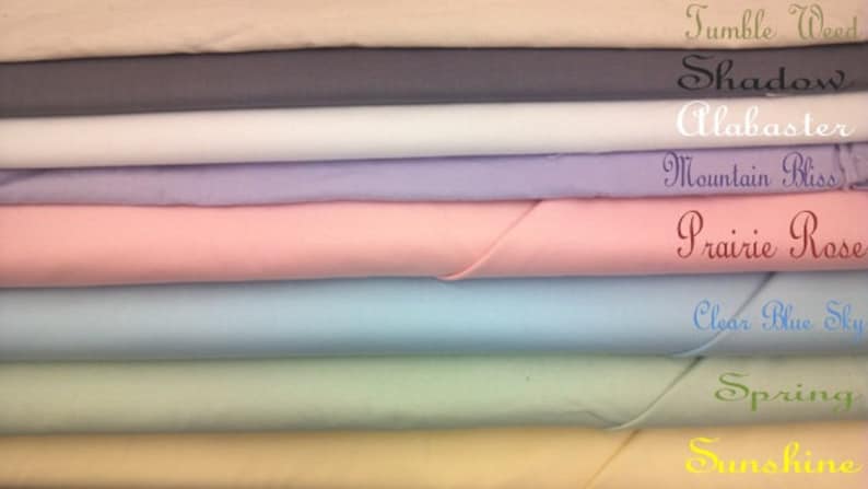 May include: A stack of eight fabric swatches in various pastel colors, including beige, gray, white, lavender, pink, light blue, green, and yellow. The swatches are labeled with names like "Tumble Weed", "Shadow", "Alabaster", "Mountain Bliss", "Prairie Rose", "Clear Blue Sky", "Spring", and "Sunshine".