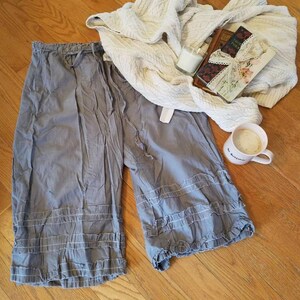May include: Gray cotton pants with a ruffled hem and a drawstring waist. The pants are laid out on a wooden floor with a white blanket, a book, and a cup of coffee.