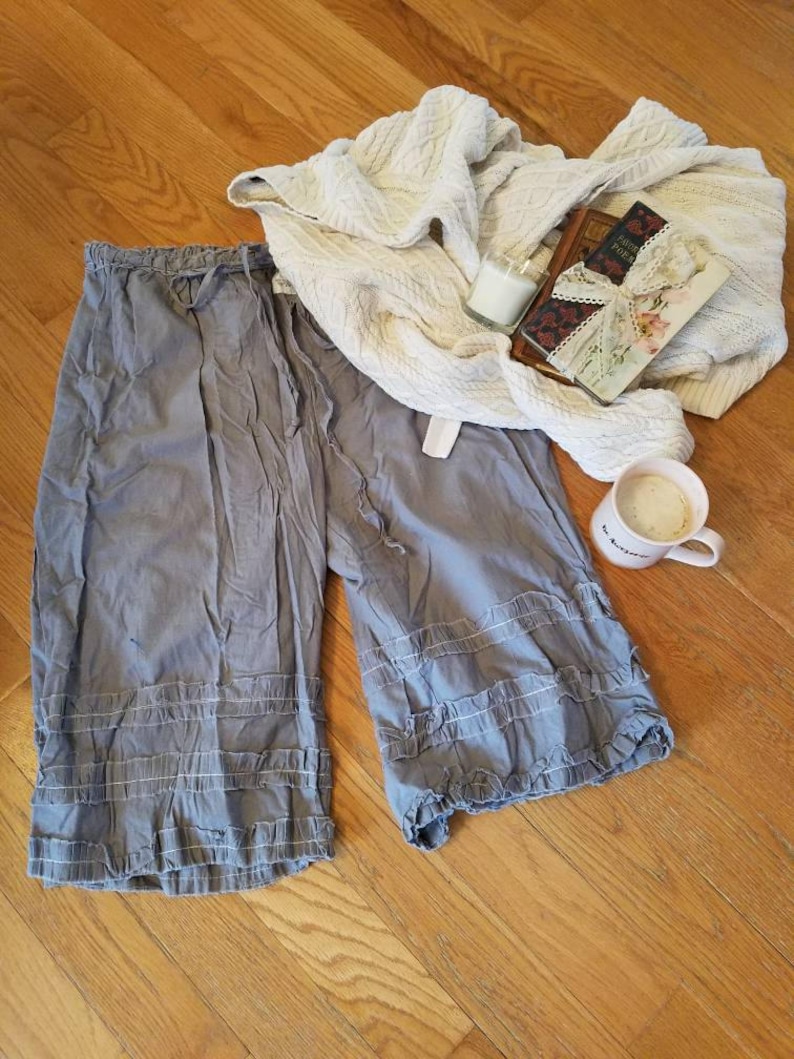 May include: Gray cotton pants with a ruffled hem and a drawstring waist. The pants are laid out on a wooden floor with a white blanket, a book, and a cup of coffee.