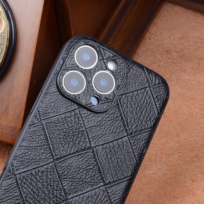 May include: Black phone case with a textured, quilted pattern. The case features a camera module with three lenses and a flash. The design is a dark, neutral color, offering a sleek and protective aesthetic.