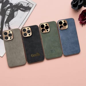 Puede incluir: Cuatro fundas para teléfono en tonos grises, negro, verde y azul. Cada funda tiene un anillo de cámara dorado y un borde dorado a juego. La funda negra tiene la palabra "Goals" en dorado, y la funda verde tiene las iniciales "TH".