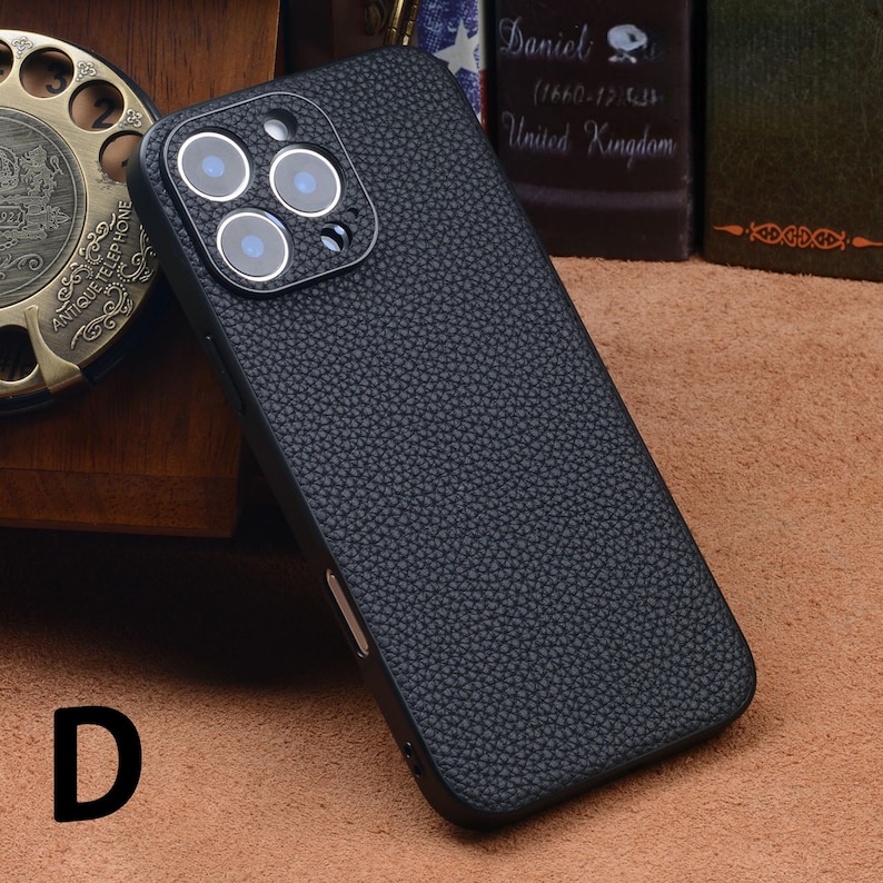 May include: A black textured phone case with a camera lens protector. The case has a leather-like appearance and is designed to fit a smartphone. The camera lens protector has three circular openings. The letter "D" is visible in the lower left corner.