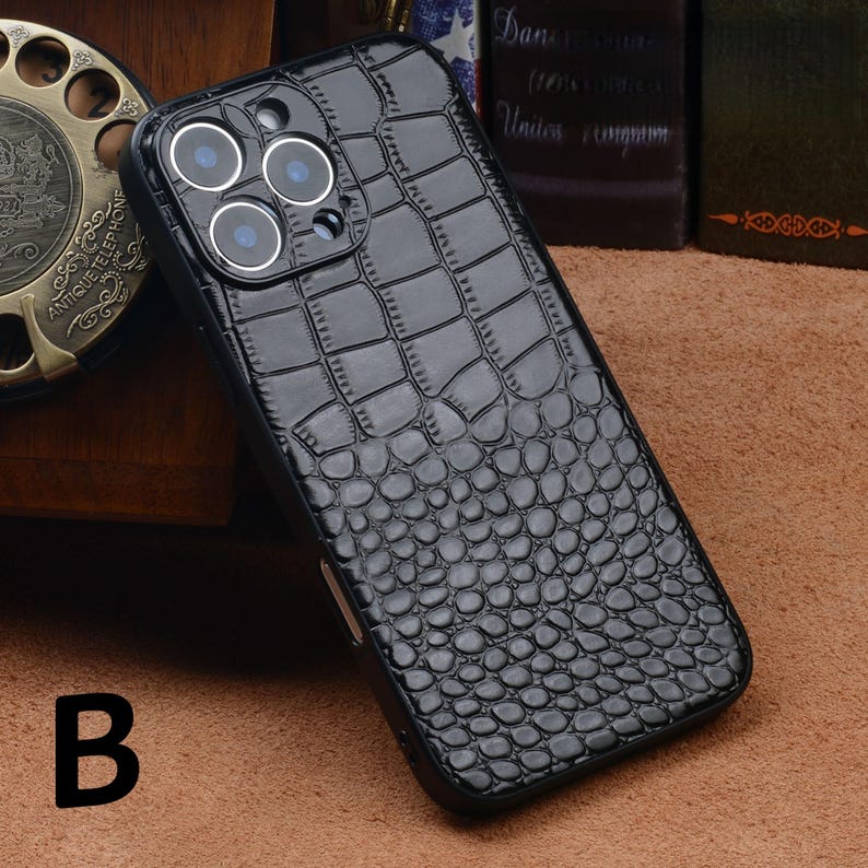 May include: Black phone case with a crocodile skin texture. The case has a glossy finish and features three camera lenses. The letter "B" is visible in the lower left corner.