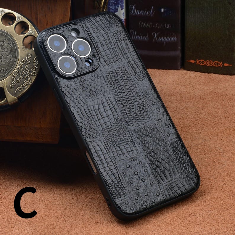 May include: Black phone case with a crocodile skin texture. The case has a camera cutout with three lenses and a flash. The letter "C" is visible in the lower left corner. The case is designed to protect the phone from scratches and damage.