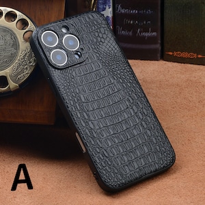 May include: Black phone case with a crocodile skin texture. The case has a camera cutout with three lenses and a flash. The letter "A" is visible in the lower left corner. The case is on a brown surface.