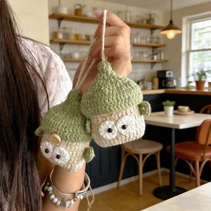 May include: Two handmade crochet owl ornaments. Each owl has a light green hat, beige body, and large white eyes with black pupils. One owl has a small wooden button. The owls are suspended by a white string.