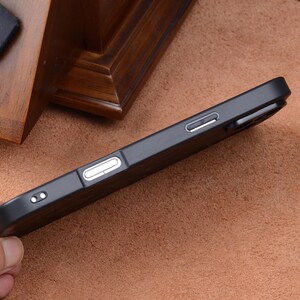 May include: A black phone case with a textured wood grain design. The case has cutouts for the phone's buttons and camera. The case is shown on a brown surface.