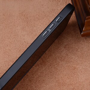 May include: A black phone case with three buttons on the side. The case is angled, showing the side profile and buttons. The case is made of a matte material and is set against a brown background.