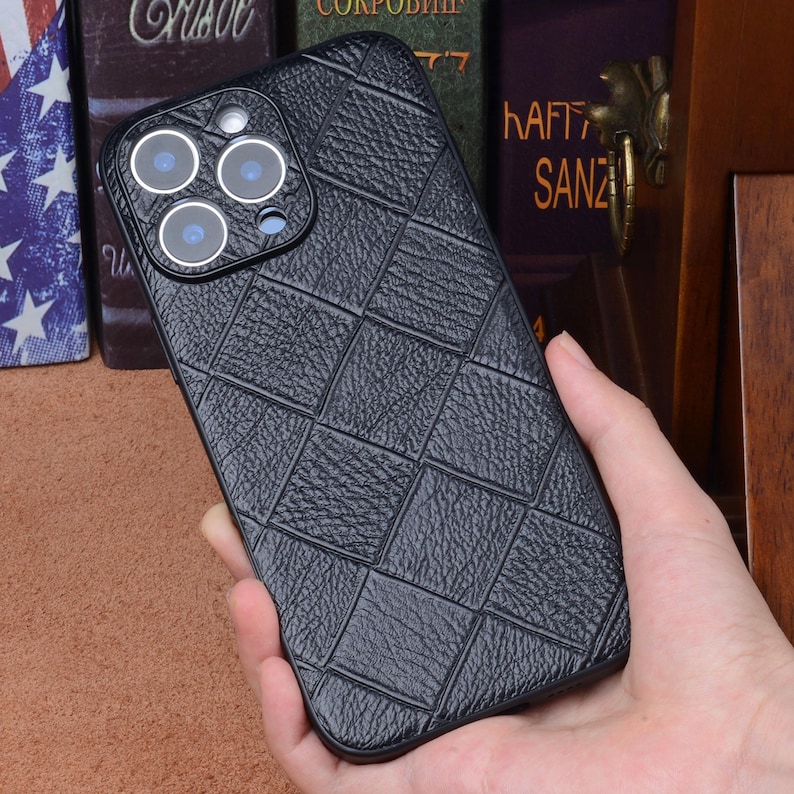 May include: A black phone case with a textured, diamond-patterned design. The case has a camera cutout with multiple lenses. The phone case is held in a person's hand.