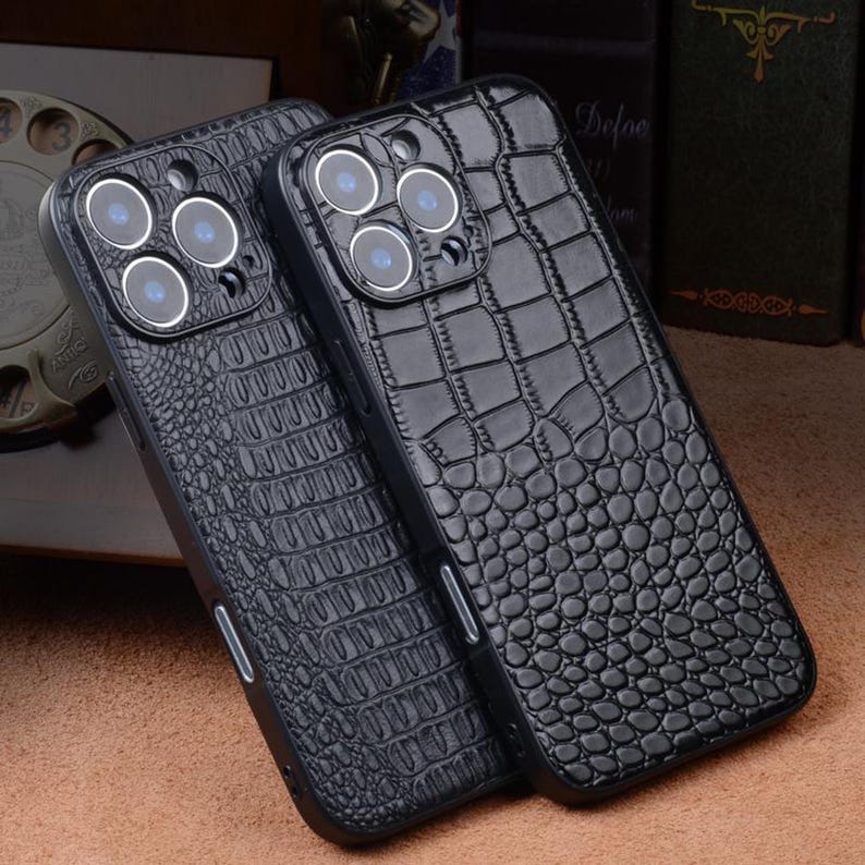 May include: Two black phone cases with a crocodile skin texture. The cases have a raised camera lens area with multiple circular lenses. The cases are made of a glossy material and are designed to protect the phone.