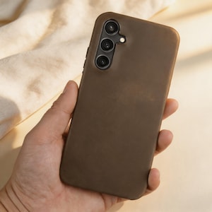 Può includere: Una custodia marrone per telefono con tre obiettivi della fotocamera è tenuta in mano. La custodia ha una finitura liscia e opaca e copre la parte posteriore e i lati del telefono. Lo sfondo è di un colore neutro.