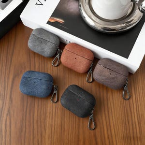 May include: Five textured leather AirPod cases in various colors: gray, brown, blue, black, and dark brown. Each case has a silver carabiner clip attached. The cases are displayed on a wooden surface.