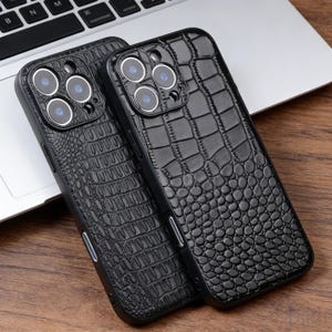 May include: Two black phone cases with a crocodile skin texture are displayed. Each case has a camera module with multiple lenses. The cases are made of a glossy material and are designed to protect the phone.