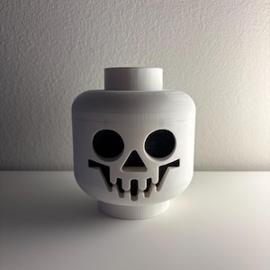 May include: A white, 3D-printed skull head, resembling a toy block head. The skull has large, black eye sockets and a detailed jawline. The head sits on a white base and has a cylindrical top. The background is a plain white wall.
