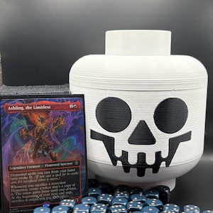May include: A white plastic container shaped like a toy head with a black skull face print, positioned next to a Magic: The Gathering card titled Ashling, the Limitless and a pile of small dark blue dice with copper-colored pips on the surface.
