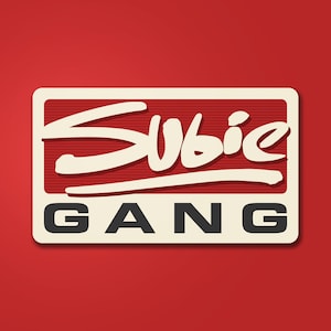 May include: A rectangular sticker with the text "Subie GANG" in a retro style. The word "Subie" is in a white, cursive font, above the word "GANG" in a black, block font. The background is red with a cream-colored border.