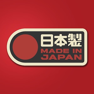 May include: A rectangular sticker with rounded edges, featuring a red circle on the left and the text "MADE IN JAPAN" in white and Japanese characters. The sticker has a black background and a cream-colored border, set against a red background.