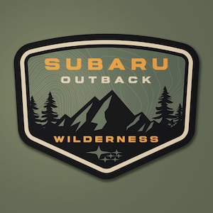 May include: A shield-shaped sticker with a black border and a green background. The sticker features the words "SUBARU OUTBACK WILDERNESS" in orange and white, along with a mountain range and pine trees graphic. The Subaru logo is at the bottom.