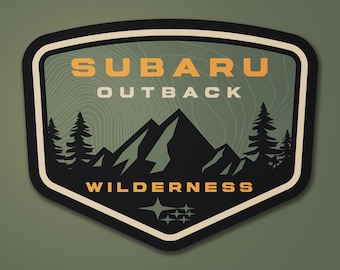 May include: A shield-shaped sticker with a black border and a green background. The sticker features the words "SUBARU OUTBACK WILDERNESS" in orange and white, along with a mountain range and pine trees graphic. The Subaru logo is at the bottom.