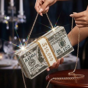 May include: A rectangular, bejeweled clutch designed to resemble a stack of $100 bills. The clutch features a gold chain strap and is embellished with sparkling rhinestones. The text "$10,000" and "OG 100" are visible on the clutch.